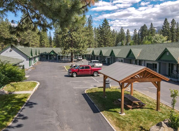 A motel with multiple green-roofed cabins, a parking lot with several vehicles, and a picnic area with benches. - Green Gables Motel & Suites (Burney)