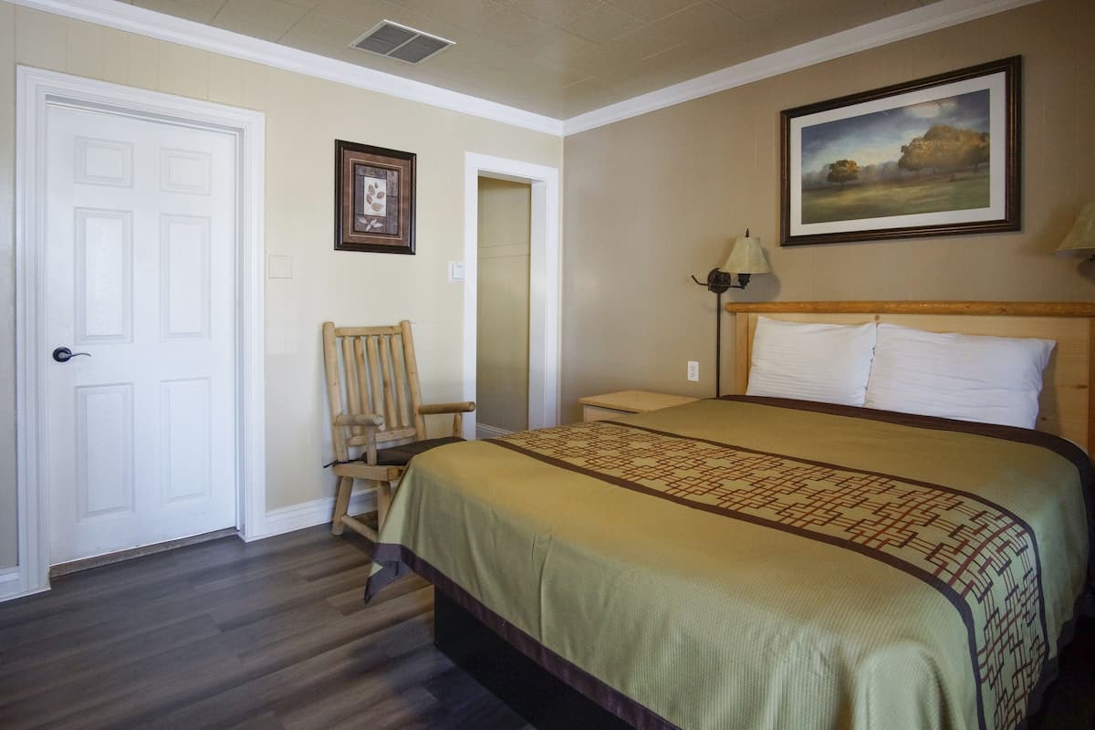 A hotel room with a bed, a chair, a door, and a painting on the wall.