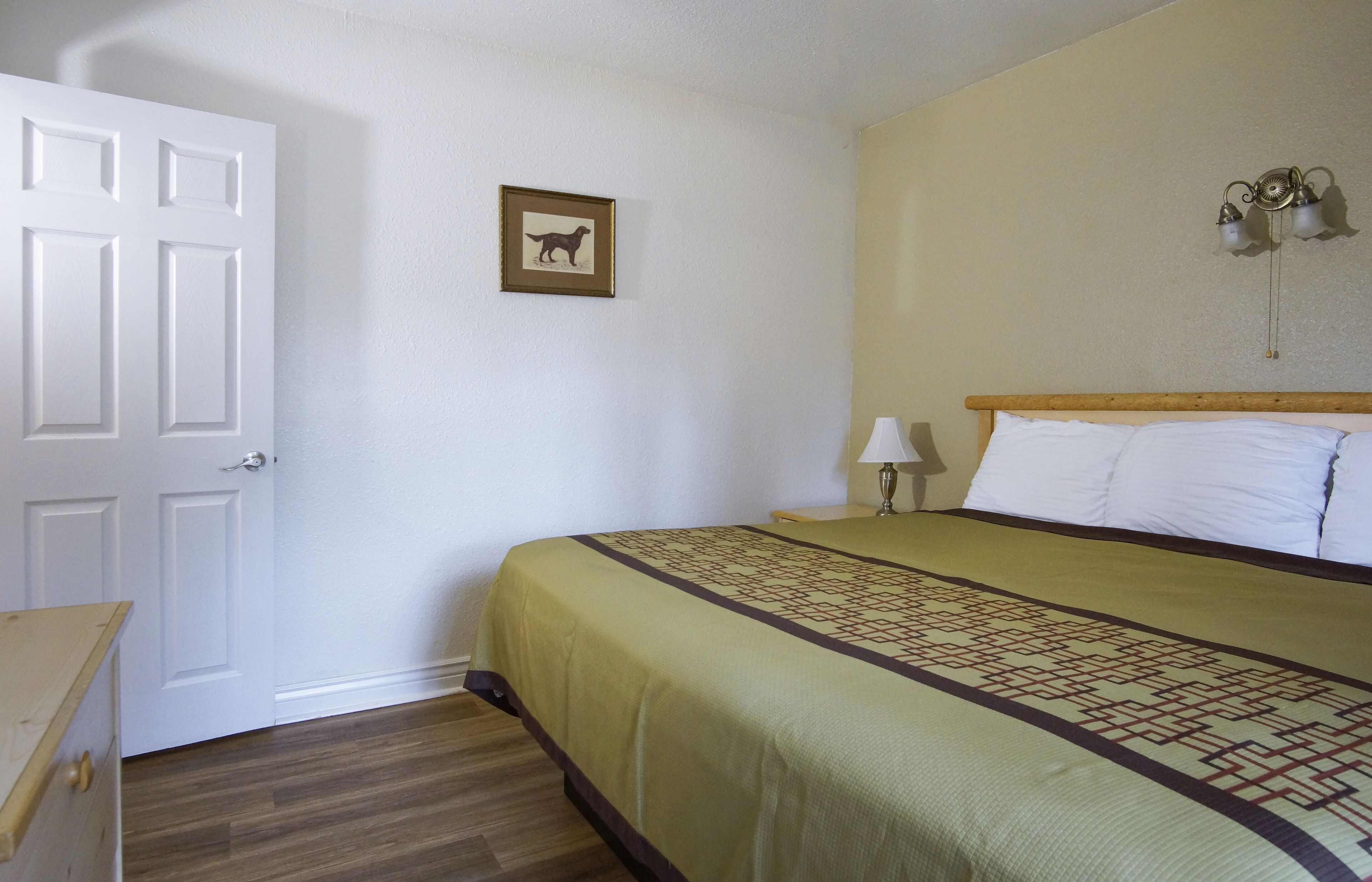 Un dormitorio con cama, una mesita de noche de madera, una lámpara de pared e imagen enmarcada.