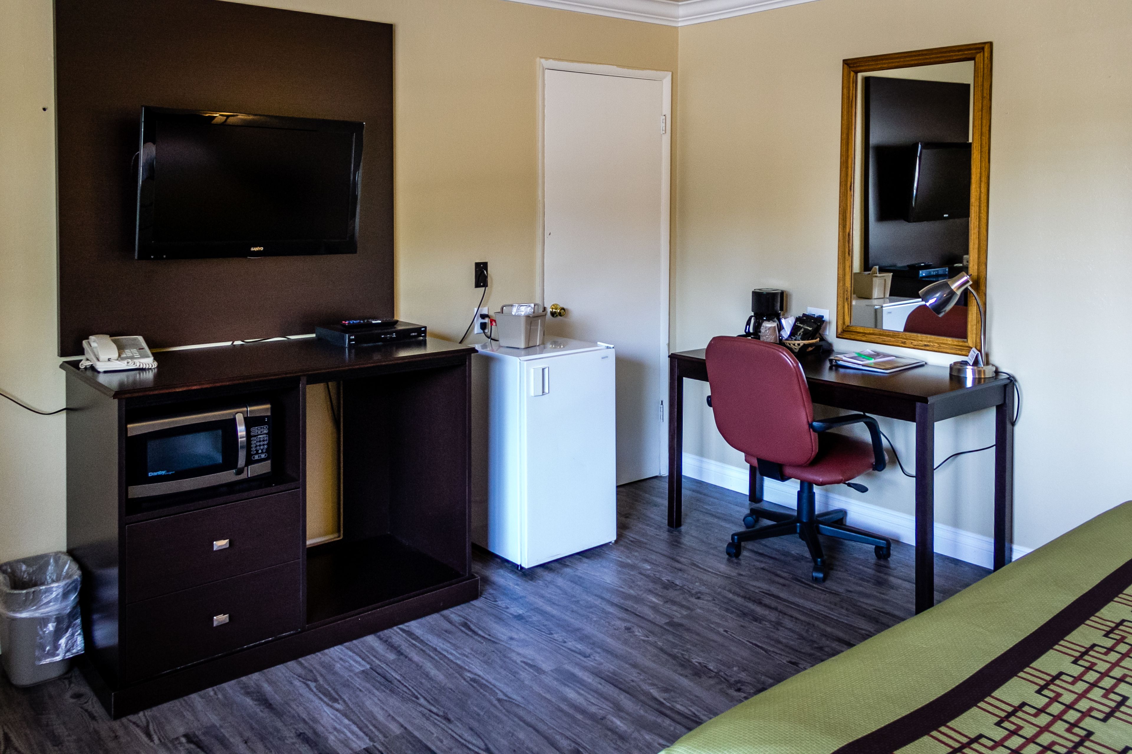 A hotel room with a dark wood desk, a flat-screen TV, a microwave, a mini-fridge, a red office chair, a desk lamp, and a large mirror.