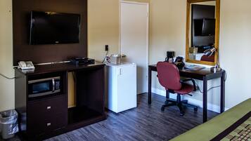 A hotel room with a dark wood desk, a flat-screen TV, a microwave, a mini-fridge, a red office chair, a desk lamp, and a large mirror.
