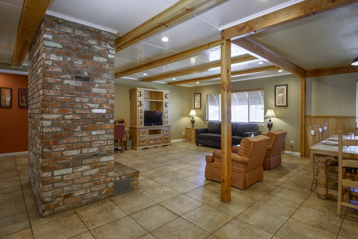 A spacious living area with a brick fireplace, wooden beams, a television, and comfortable seating.