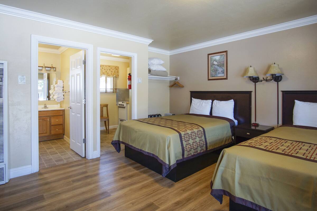 A hotel room with two beds, wooden flooring, a bathroom with a shower, and a hallway leading to another room.