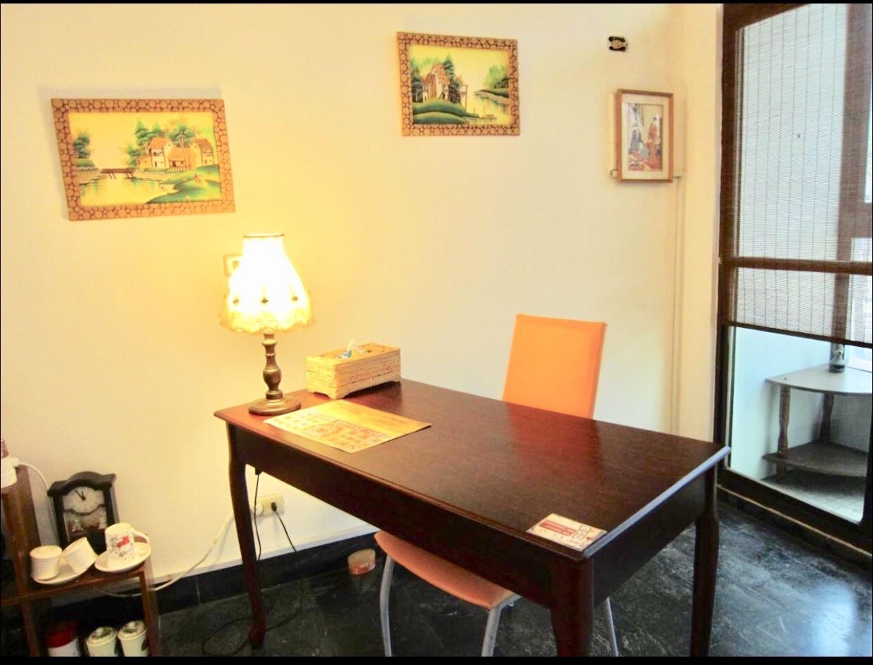 A small, simple room with a wooden table, a single chair, a lamp, and a few framed pictures on the wall.