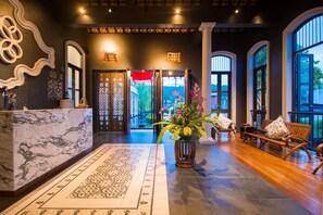 A lobby with a marble reception desk, patterned carpet, wooden benches, and large windows with a view of greenery. - Little Nyonya Hotel (Wichit)