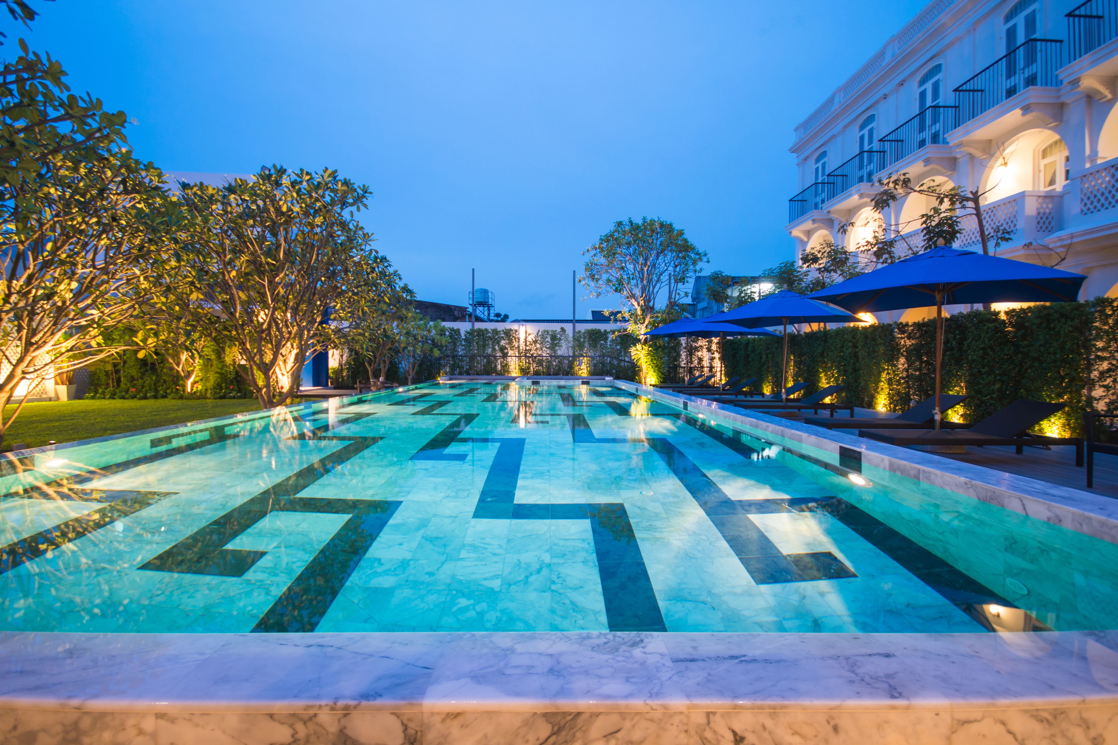 A swimming pool with geometric tile patterns, surrounded by lounge chairs and umbrellas, and adjacent to a building with balconies.