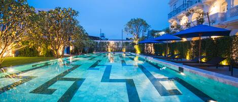 A swimming pool with geometric tile patterns, surrounded by lounge chairs and umbrellas, and adjacent to a building with balconies.