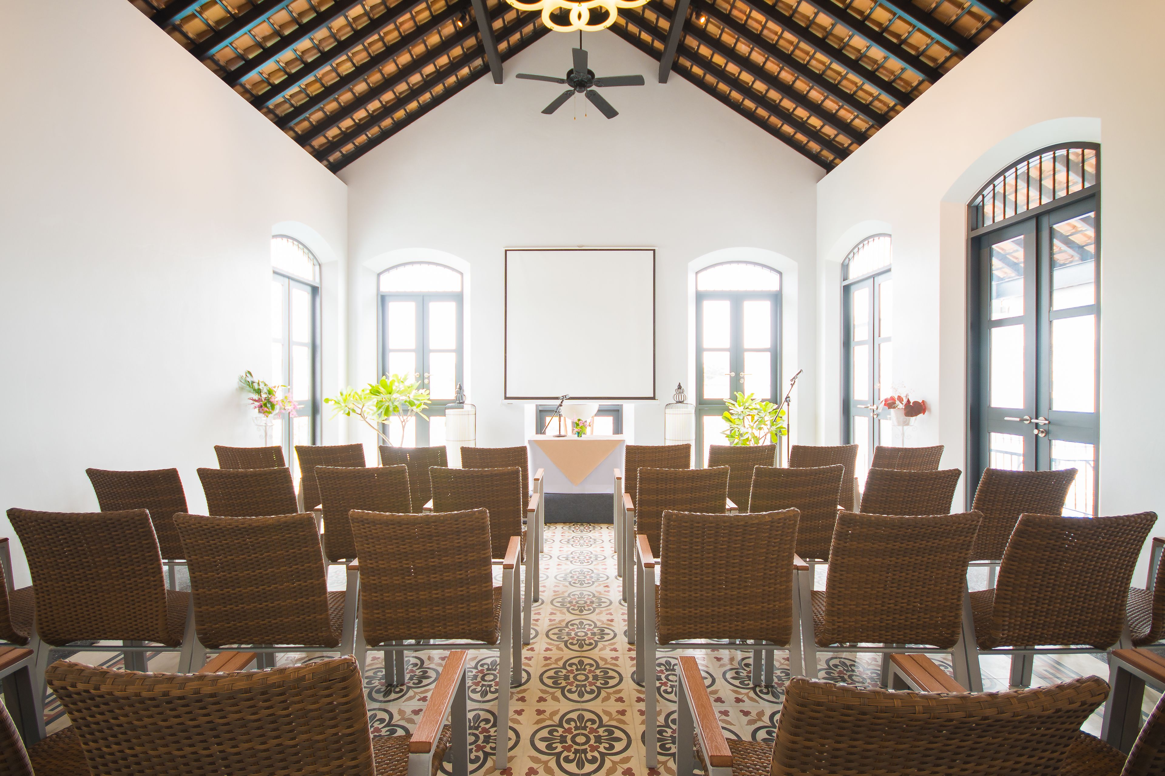 A conference room with wicker chairs arranged in rows, a central table, and a ceiling fan.