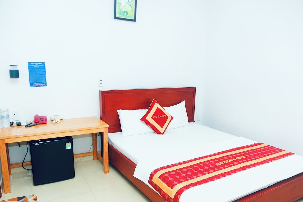 A hotel room with a wooden desk, a compact refrigerator, a bed with a red and white bedspread, and a red pillow with text.