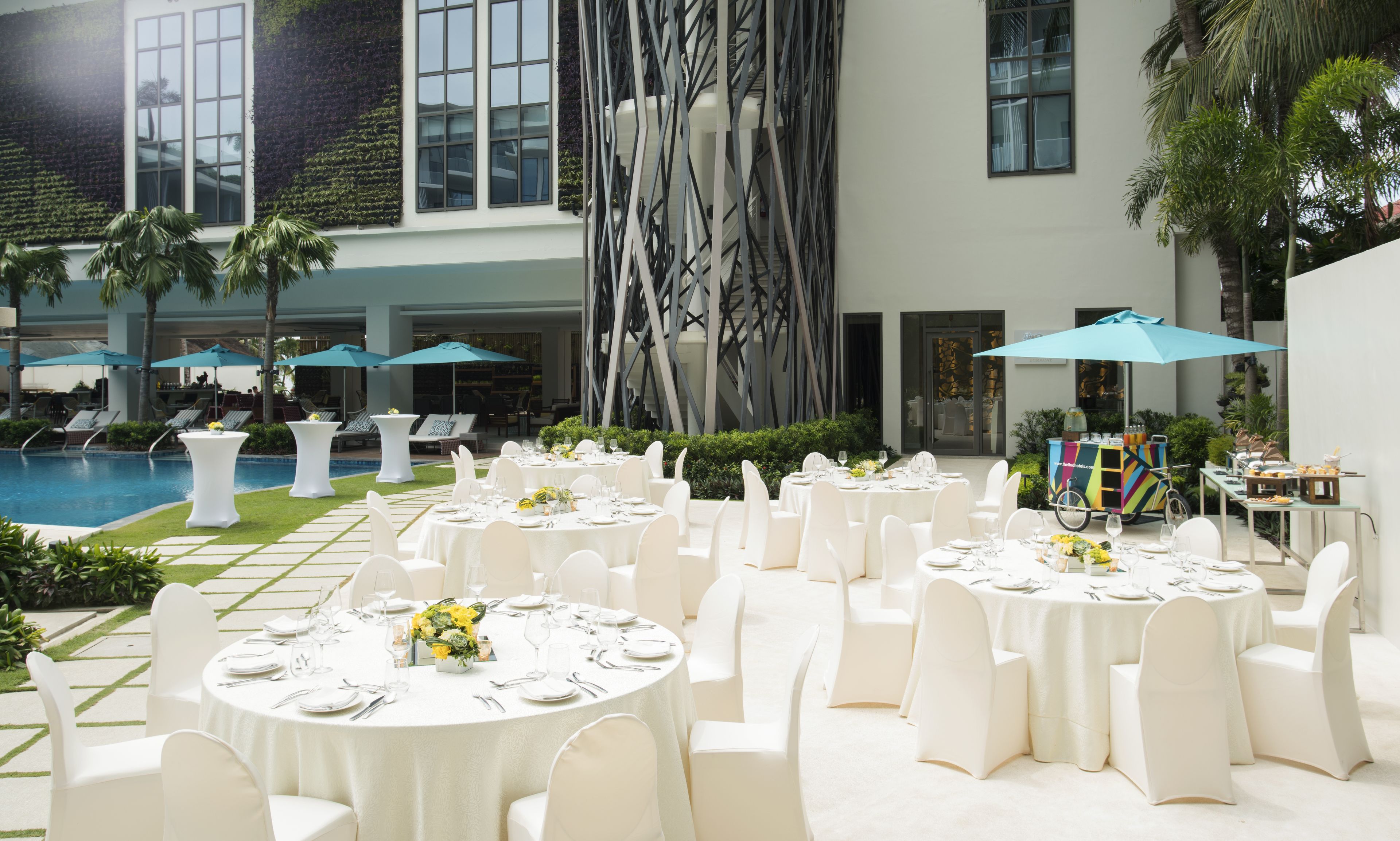 Un área de comedor exterior formal con mesas redondas, sillas blancas y sombrillas.