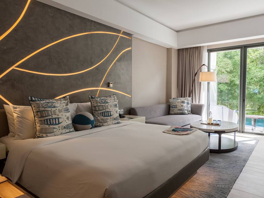 A modern hotel room with a large bed, a sofa, a small table, and a view of a pool.