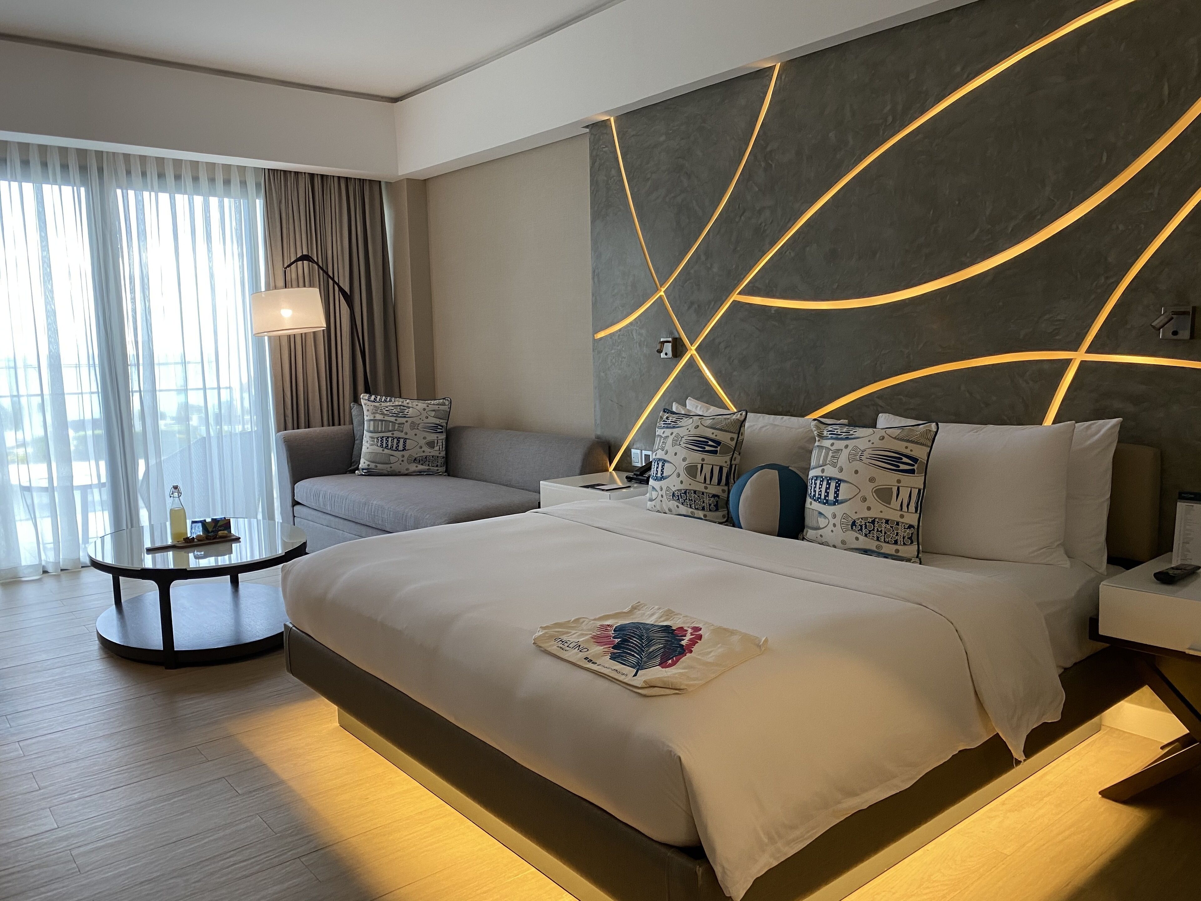 Una habitación de hotel moderna con una cama grande, un sofá, una mesa redonda y vistas al exterior a través de cortinas translúcidas.