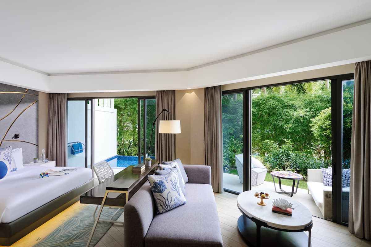 A modern hotel room with a bed, sofa, and a small table. It has large windows offering a view of greenery outside.
