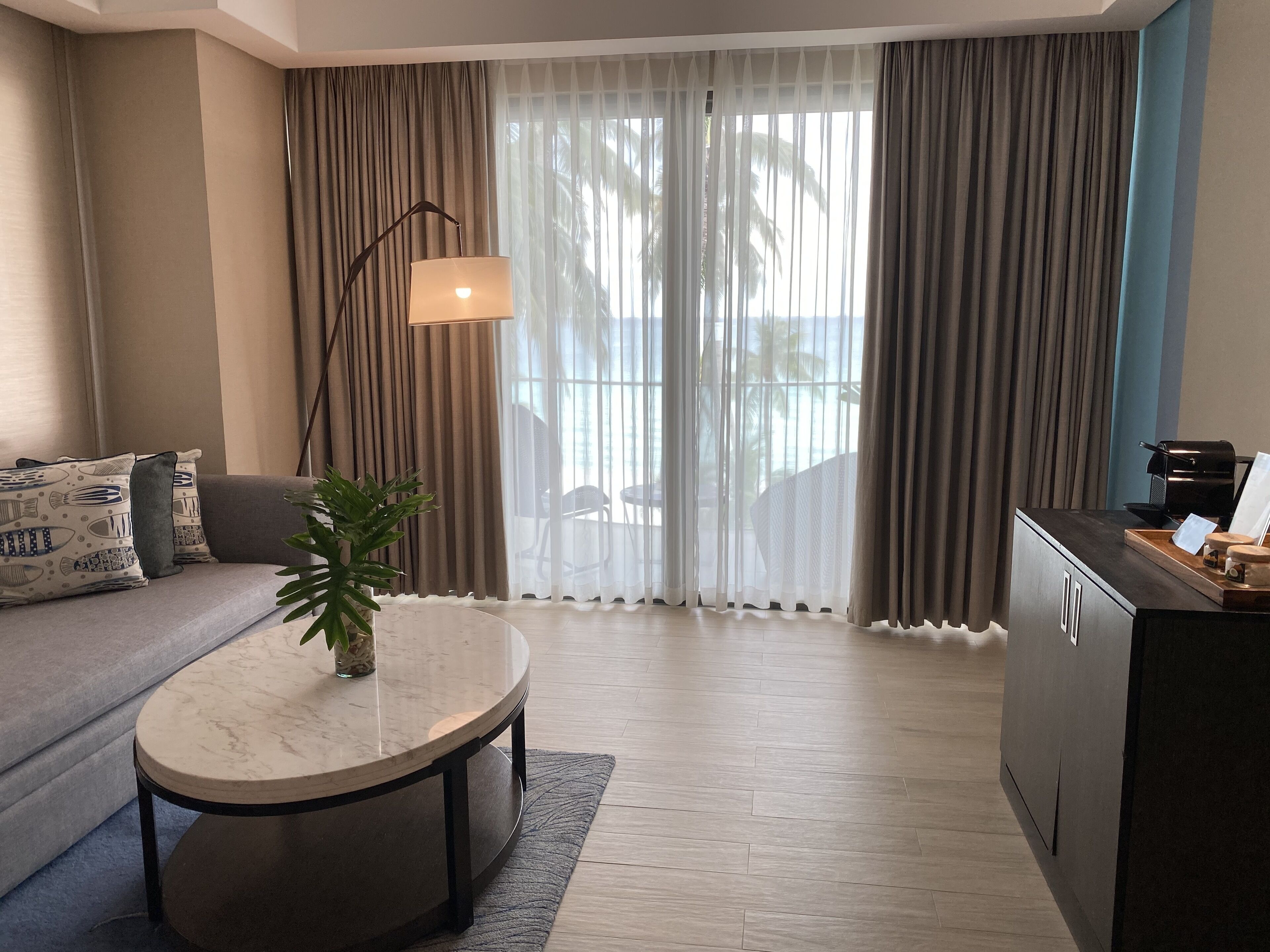 En moderne stue med sofa, rundt sofabord, gulvlampe og udsigt gennem store vinduer med gardiner.