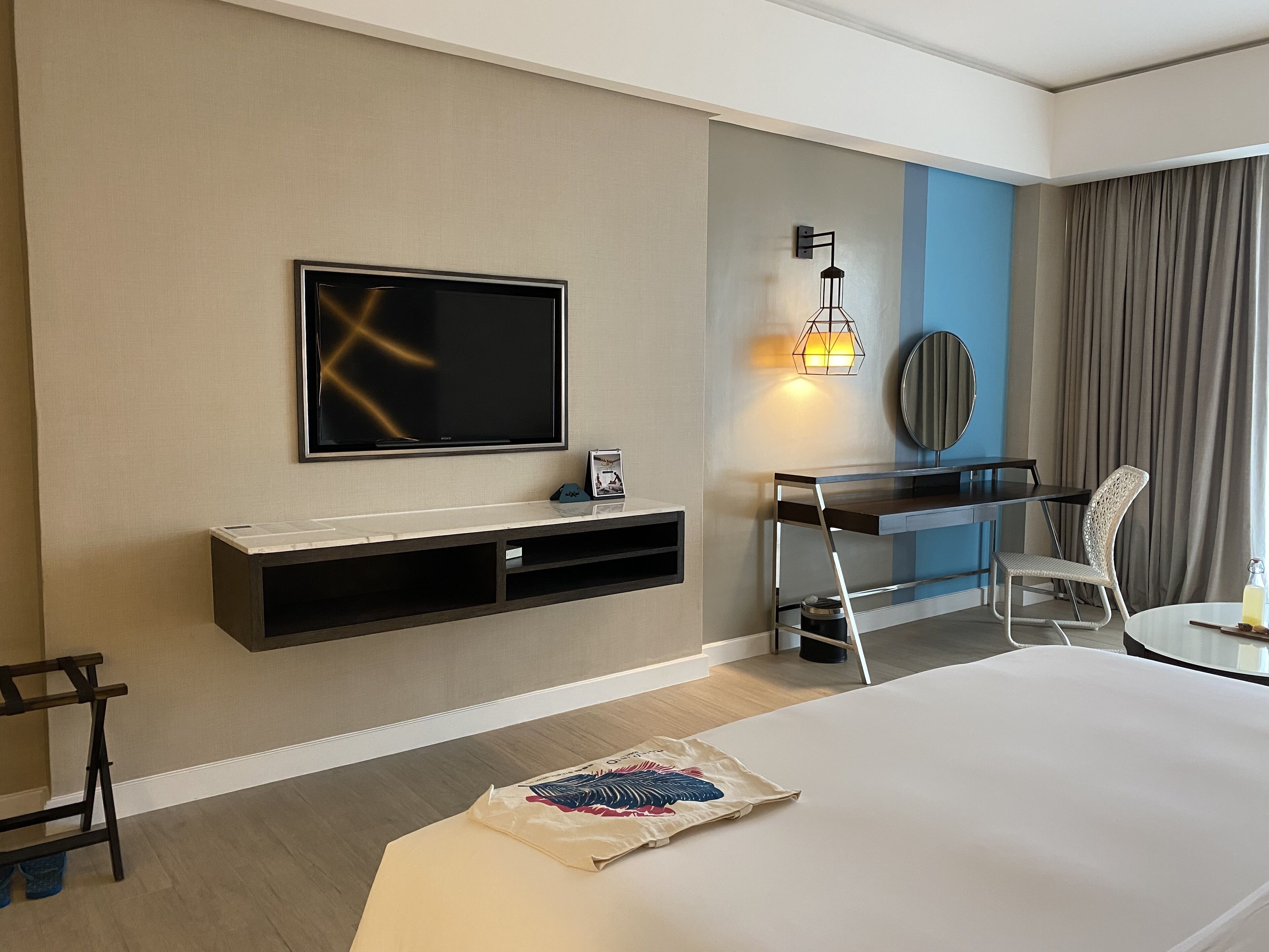 A hotel room with a flat-screen TV mounted on the wall, a desk with a chair, a bedside table, and a bed.