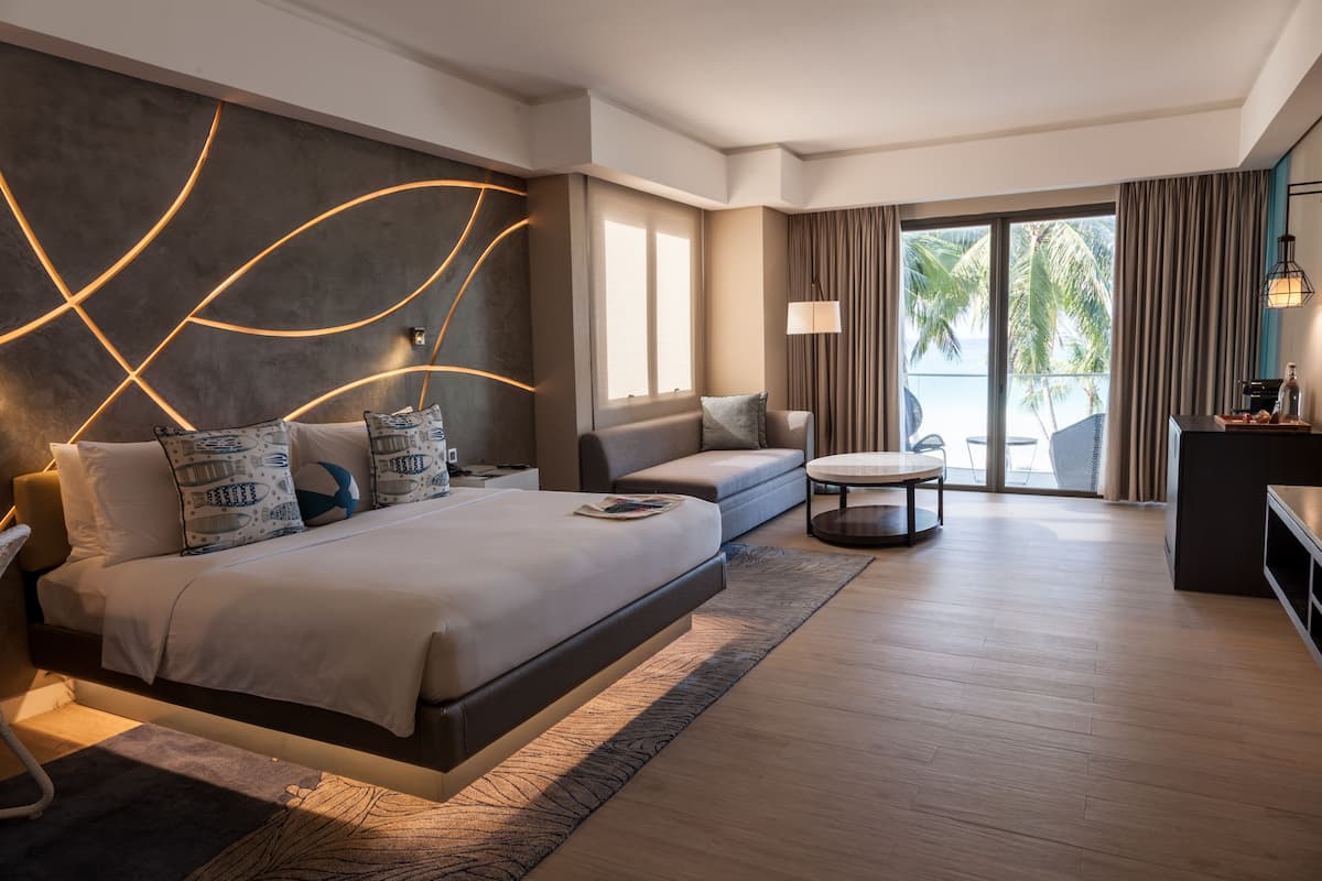A modern hotel room with a large bed, a sofa, a small table, and a balcony with a view of palm trees.