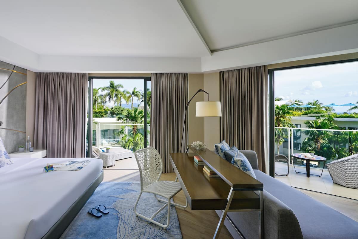 A modern hotel room with a bed, a desk, and a balcony with a view of palm trees and a pool.
