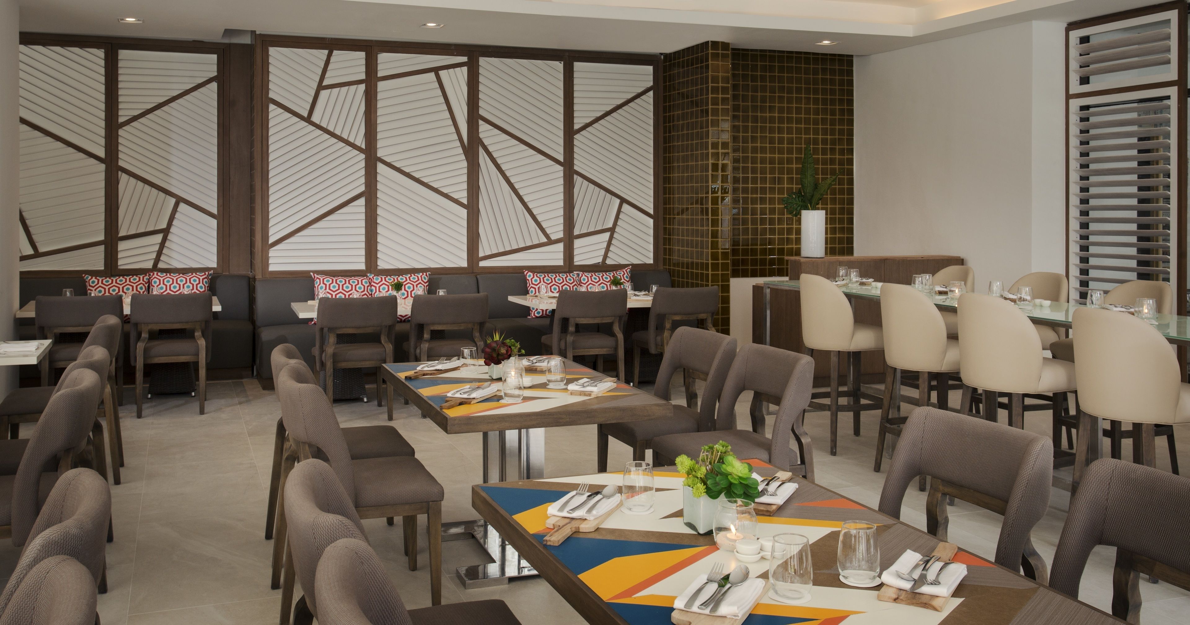 A modern dining area with multiple tables and chairs, geometric patterned walls, and a mix of natural and artificial lighting.