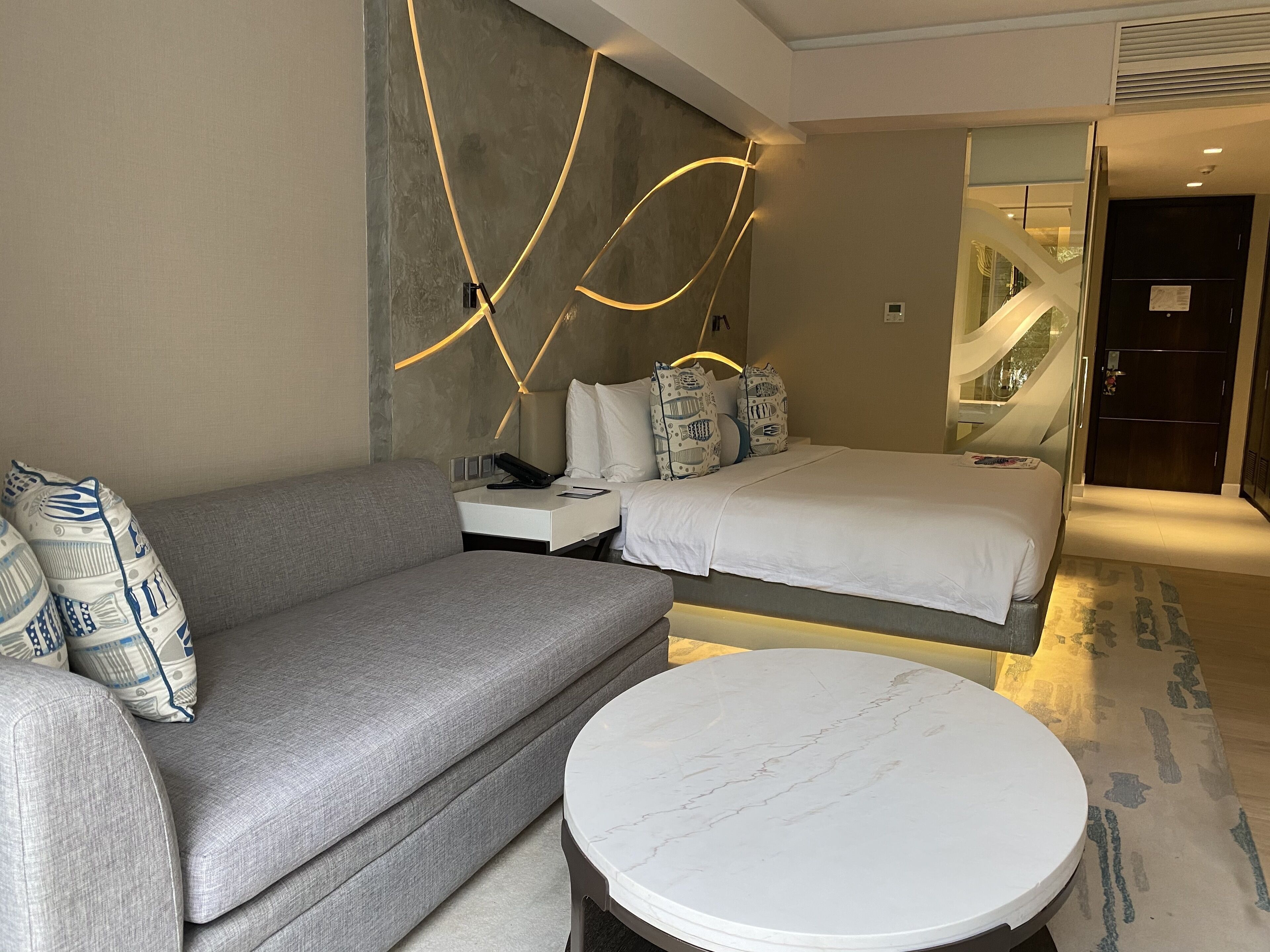 A modern hotel room with a grey sofa, a bed with patterned pillows, a bedside table with a phone, and a marble round table.