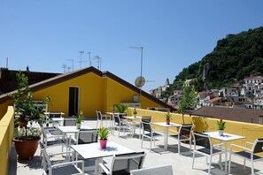 Eine Dachterrasse mit weißen Tischen und Stühlen, Kübelpflanzen und einem gelben Gebäude mit Satellitenschüssel.