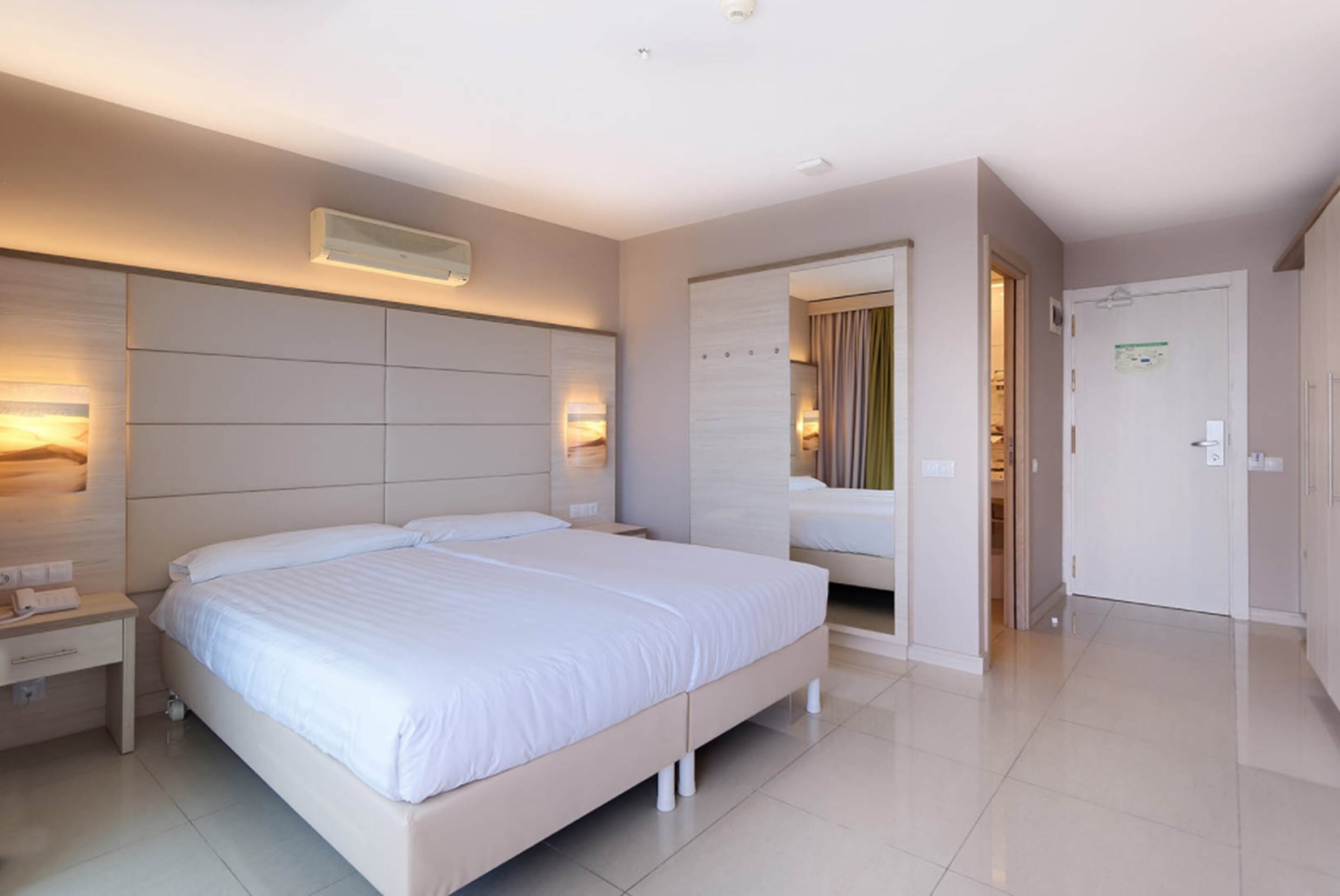 A modern hotel room with a large bed, bedside tables, an air conditioning unit, and a door leading to a bathroom.