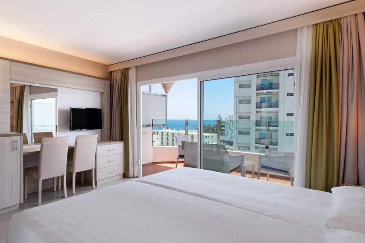 Een hotelkamer met een bed, een bureau met stoelen, een televisie en een balkon met uitzicht op gebouwen en de zee.