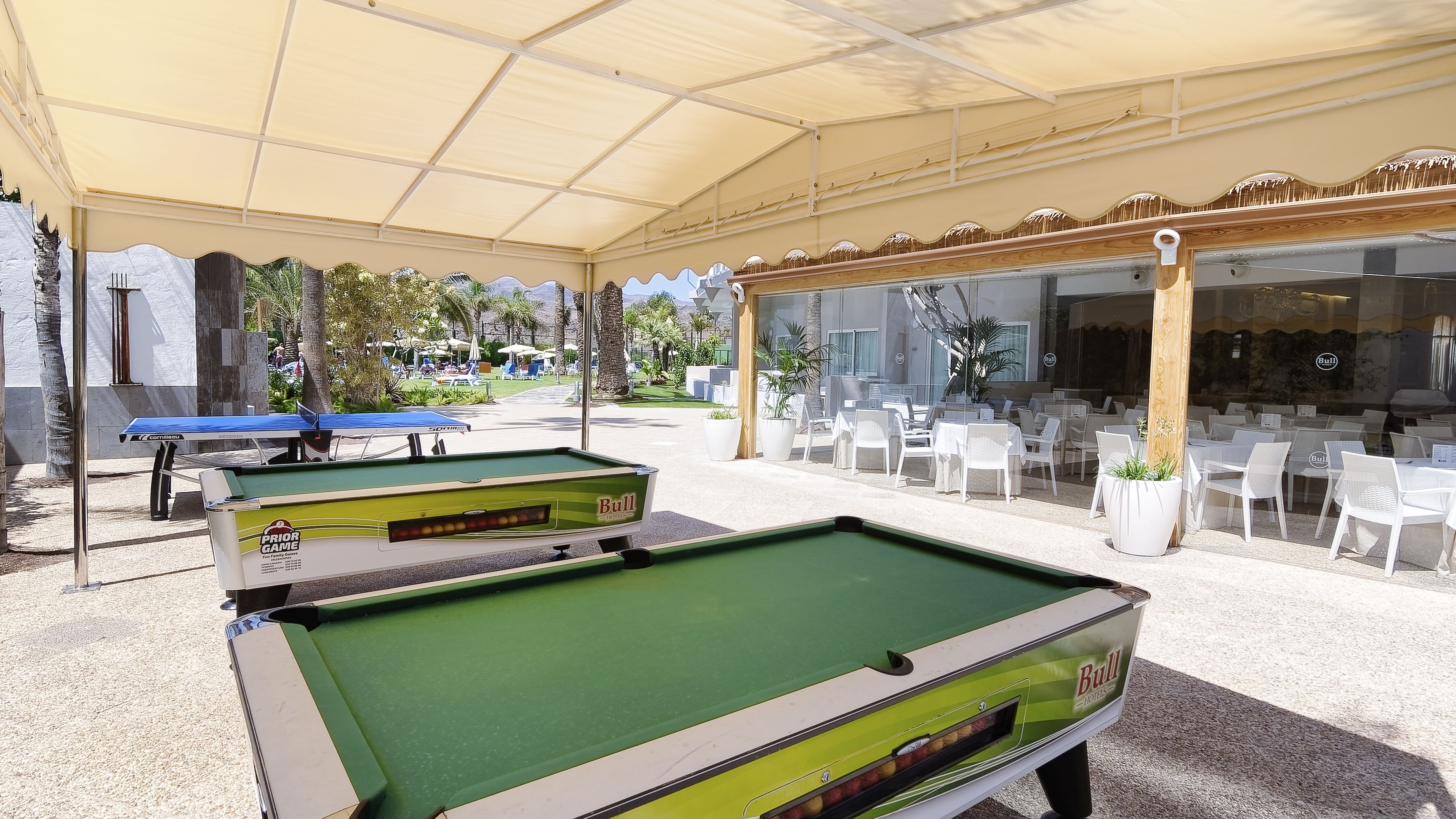 An outdoor pool area with two pool tables, a covered seating area, and a Bull Hotels restaurant in the background.
