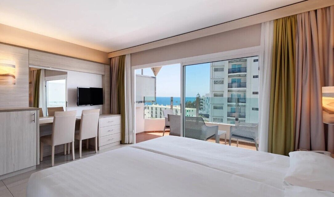 Una habitación de hotel con cama, escritorio con sillas, televisor y balcón con vista a edificios y al mar.