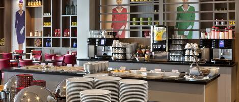 A buffet area with multiple food stations, including a coffee station and a salad bar. There are stacks of white plates and bowls, and a variety of condiments and beverages are available.