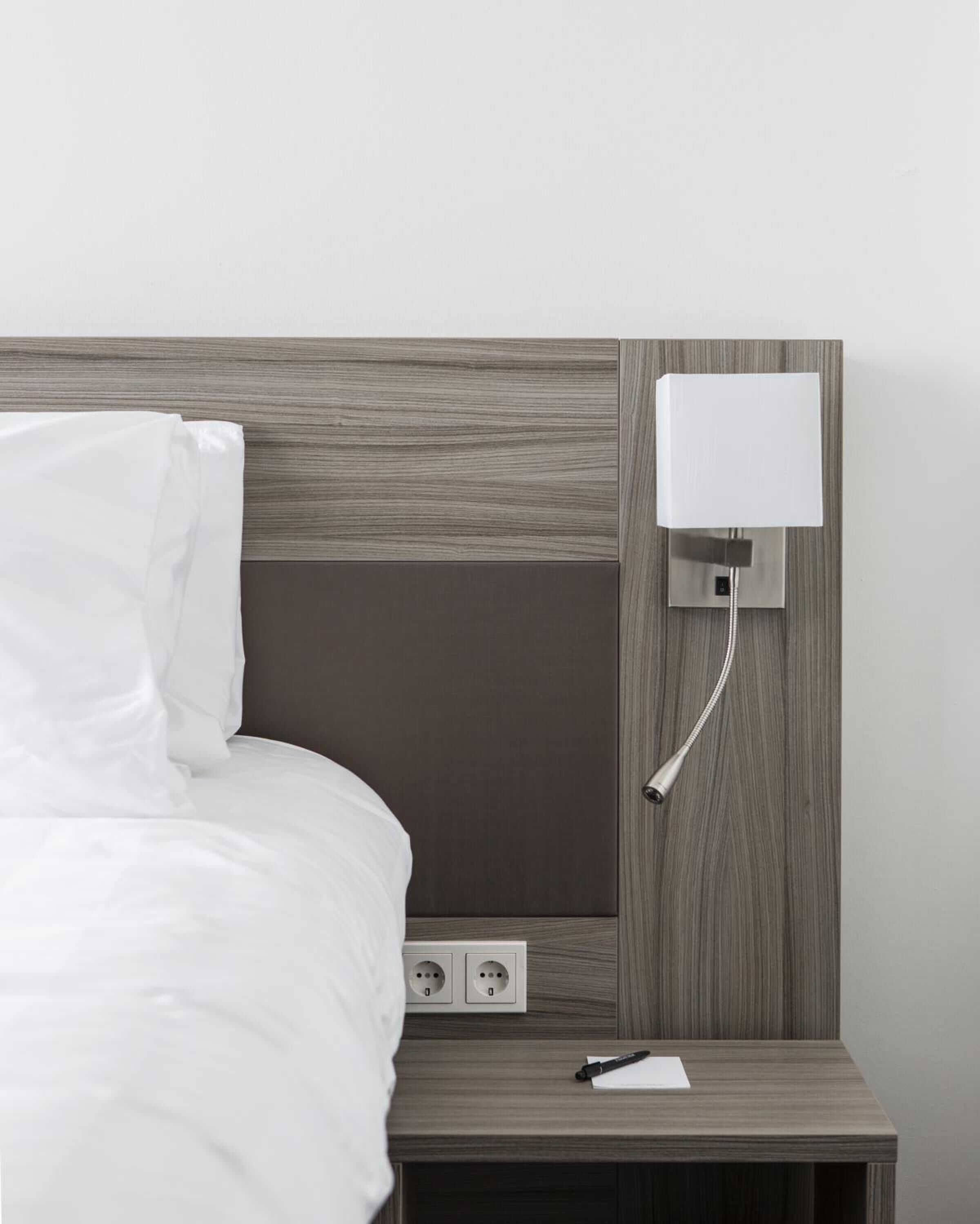Una habitación de hotel moderna con un cabecero de madera, una mesita de noche con bolígrafo y papel, y una luz nocturna en la pared.