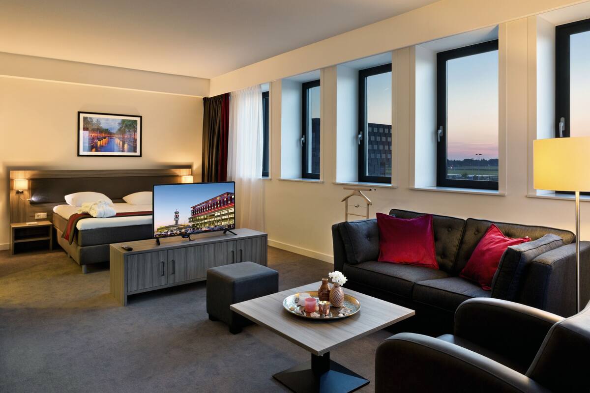 Ein Hotelzimmer mit Bett, Fernseher, Sofa, Couchtisch und Fenster mit Blick auf die Stadt.