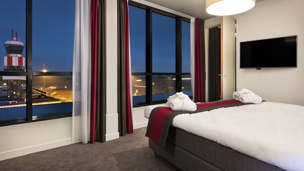 Une chambre d’hôtel avec une grande fenêtre, un lit, une télévision et une vue sur un aéroport.
