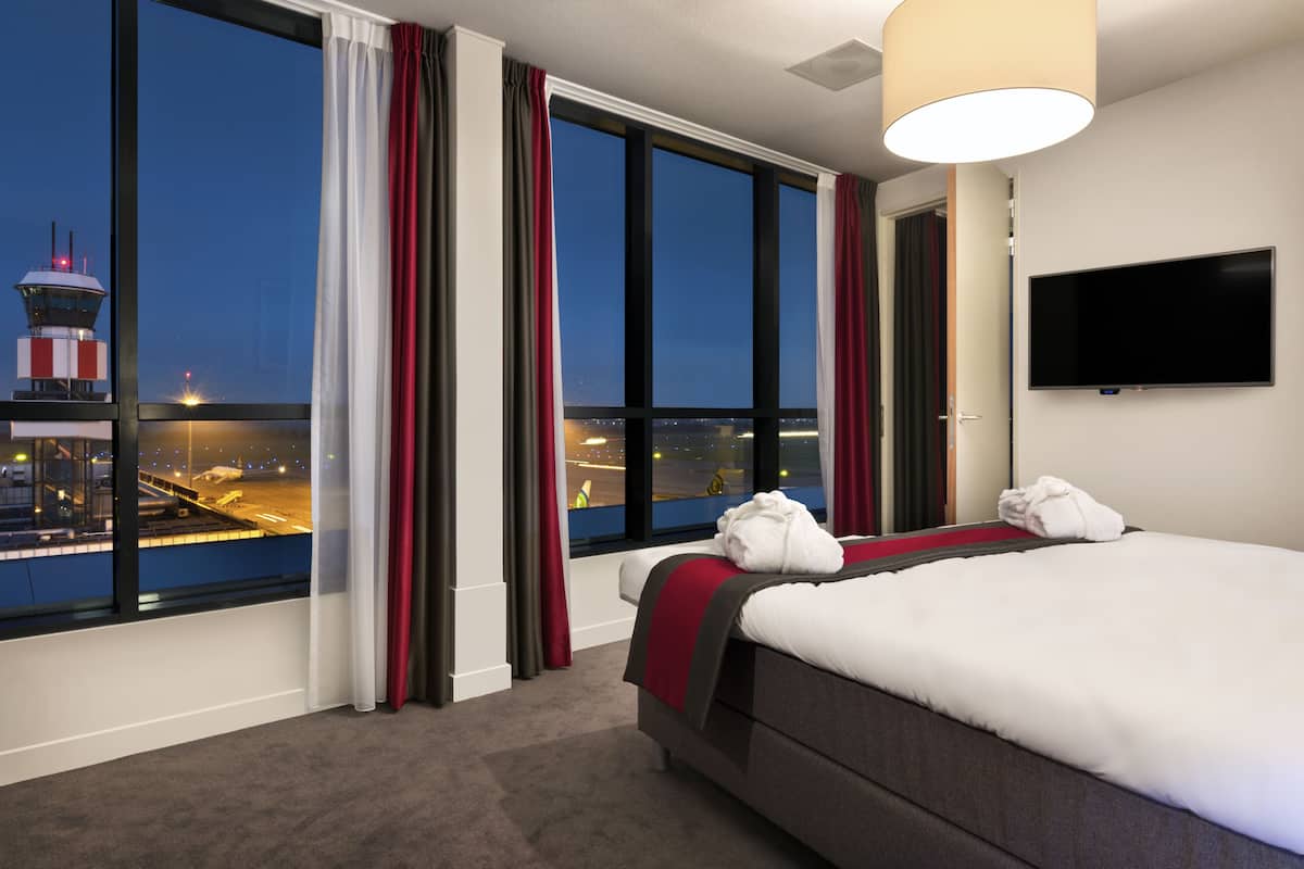Ein Hotelzimmer mit großem Fenster, einem Bett, einem Fernseher und Blick auf einen Flughafen.