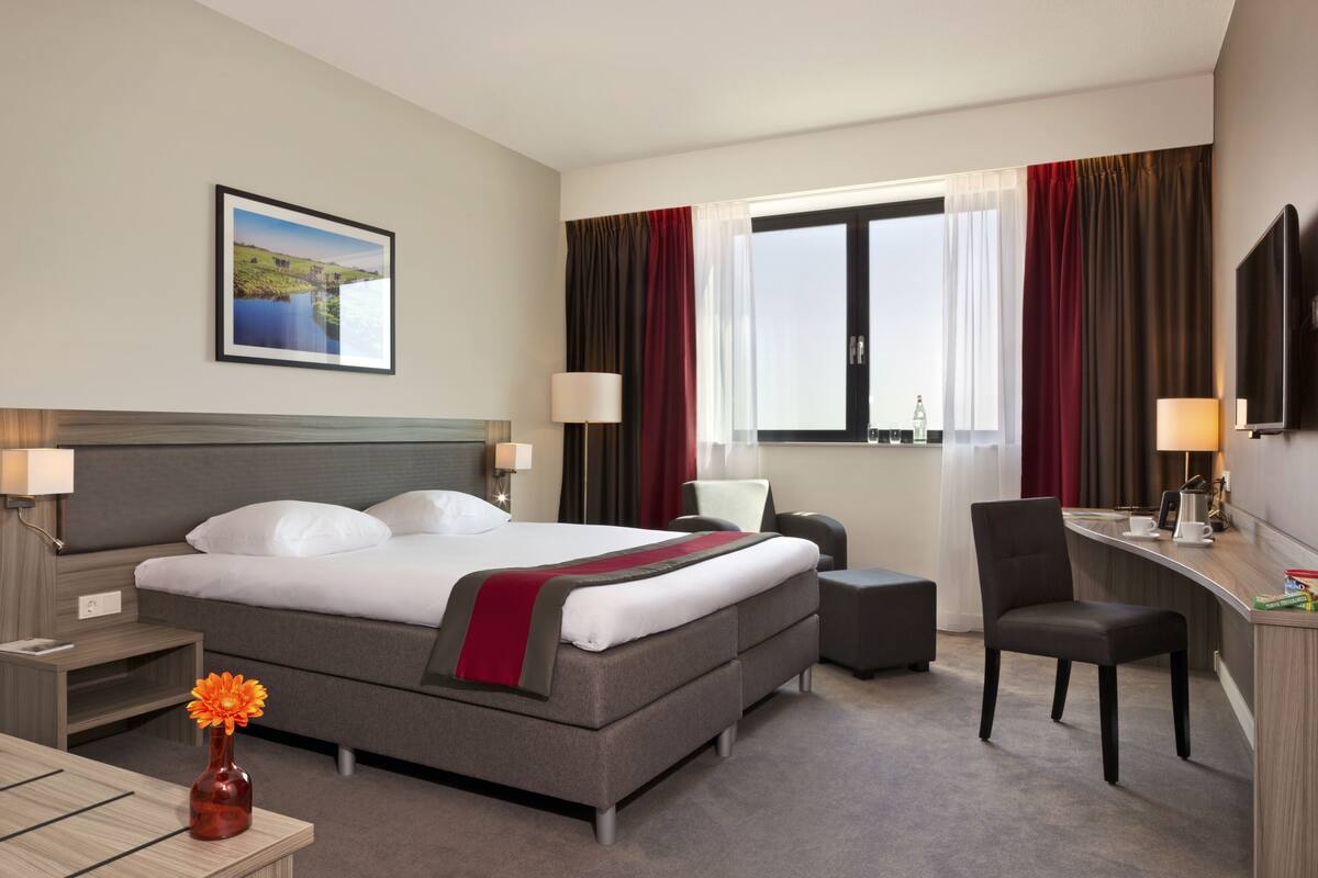 Ein Hotelzimmer mit Bett, Schreibtisch, Stuhl, Nachttisch, Lampe, einer Vase mit Blume und einem Bild an der Wand.