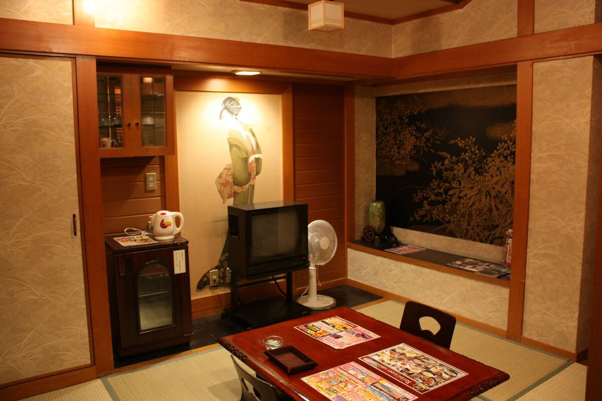 A traditional Japanese-style room with tatami flooring, a television, a small cabinet with a tea set, and a large window with a view of trees.