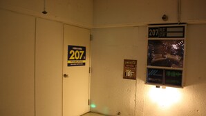 Een hotelkamer met een deur waarop '207' staat, een bordje met de tekst '207 Rustverblijf' en een poster met Japanse tekst.