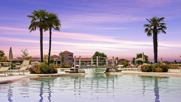 A pool area with lounge chairs, palm trees, and a building in the background. - Chervò Residence San Vigilio (Pozzolengo)