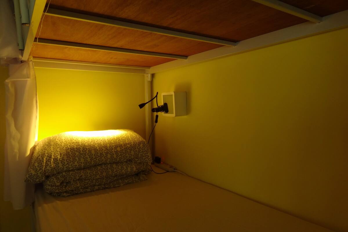 A small, dimly lit room with a single bed covered in patterned bedding, a wall-mounted light, and a power outlet.