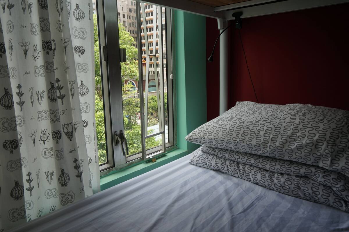 A room with a bed, a window with curtains, and a view of buildings outside.