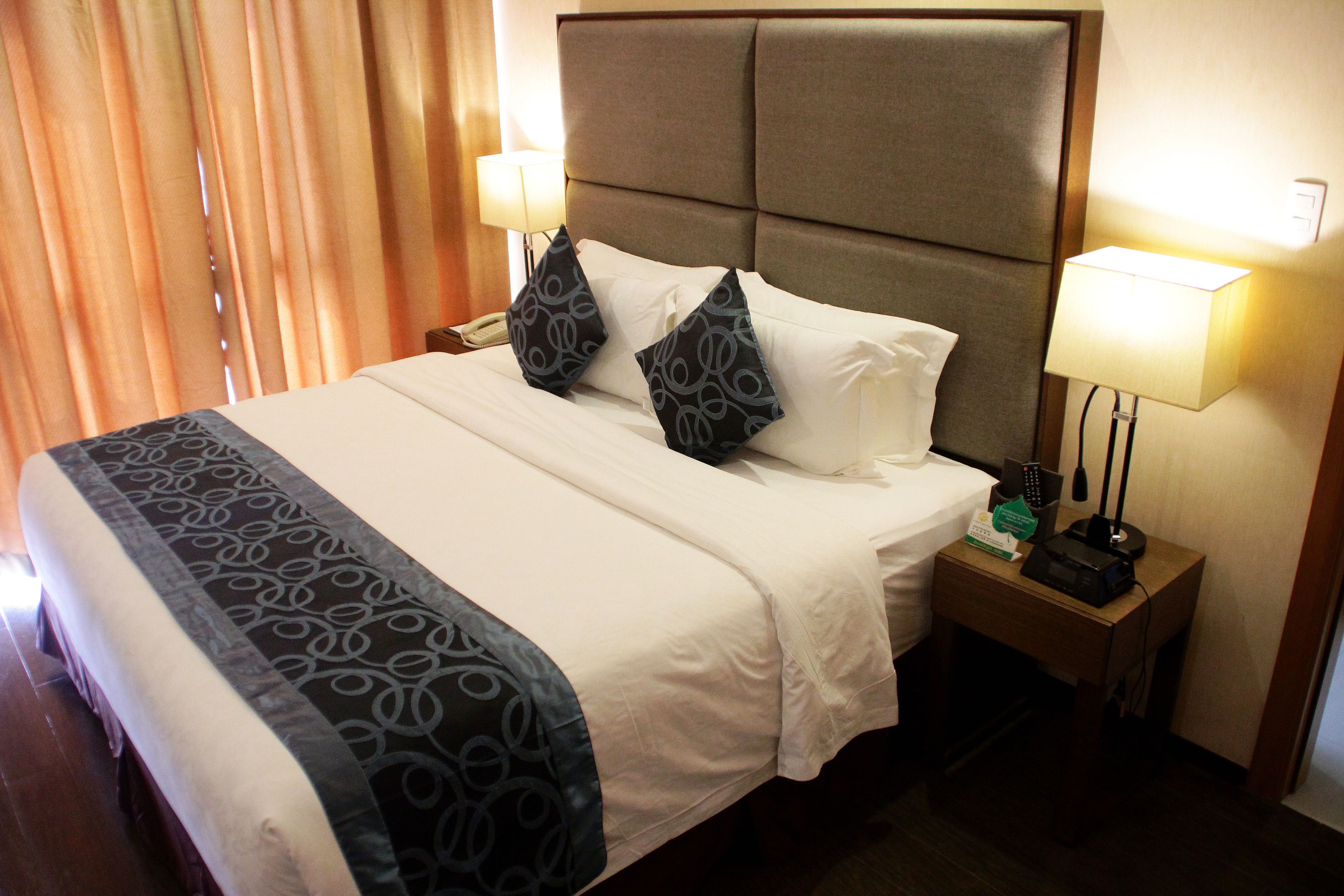 A hotel room with a large bed, two bedside lamps, a nightstand with a phone and a remote control, and a window with curtains.