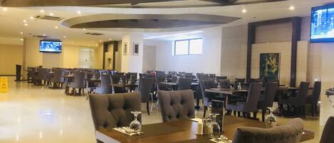 A spacious dining area with multiple tables and chairs, a flat-screen TV mounted on the wall, and a modern ceiling design.