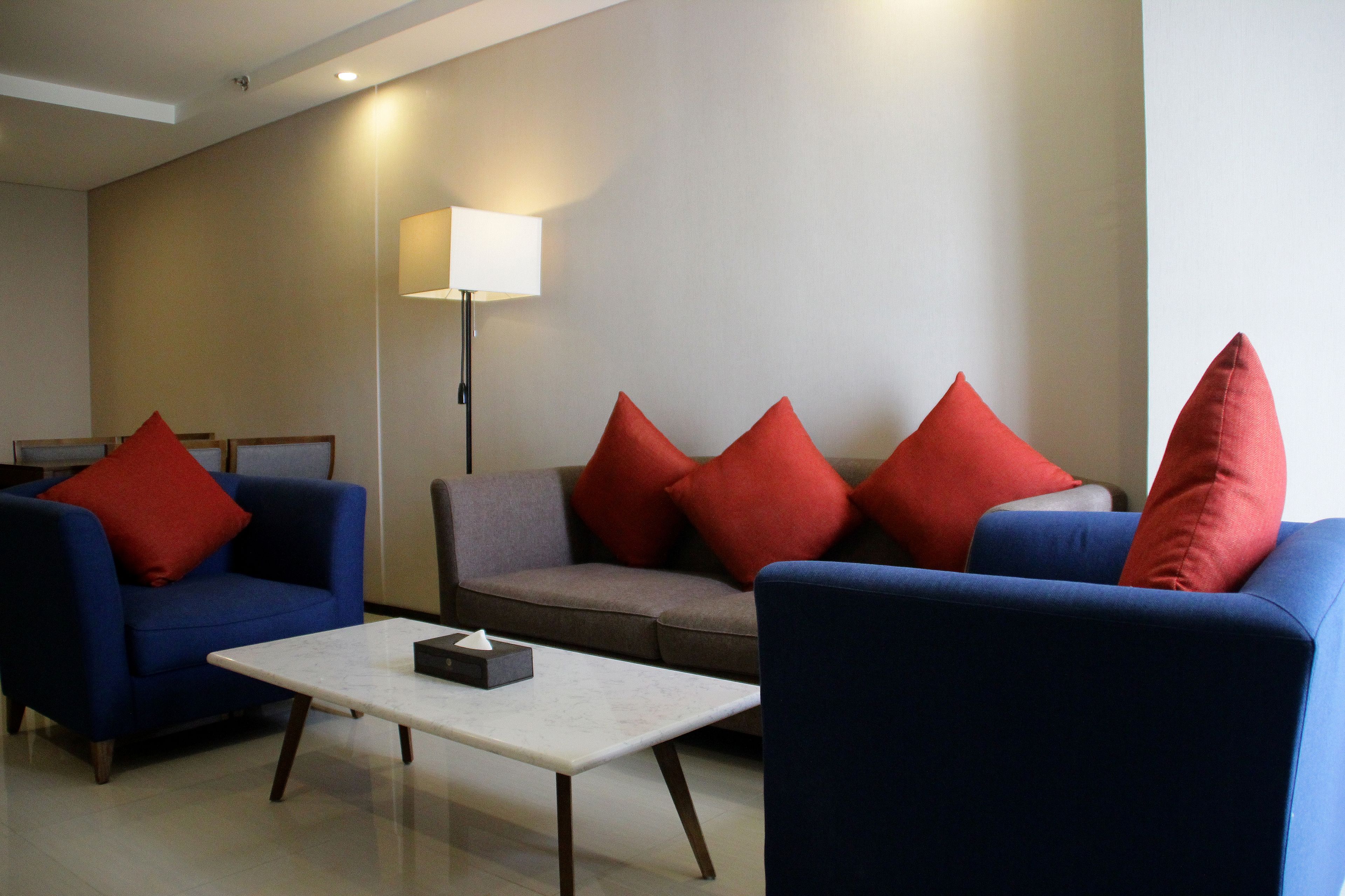 A modern living room with a blue sofa, a grey sofa, and a blue armchair, each with red cushions. A white table with a tissue box is in the center.