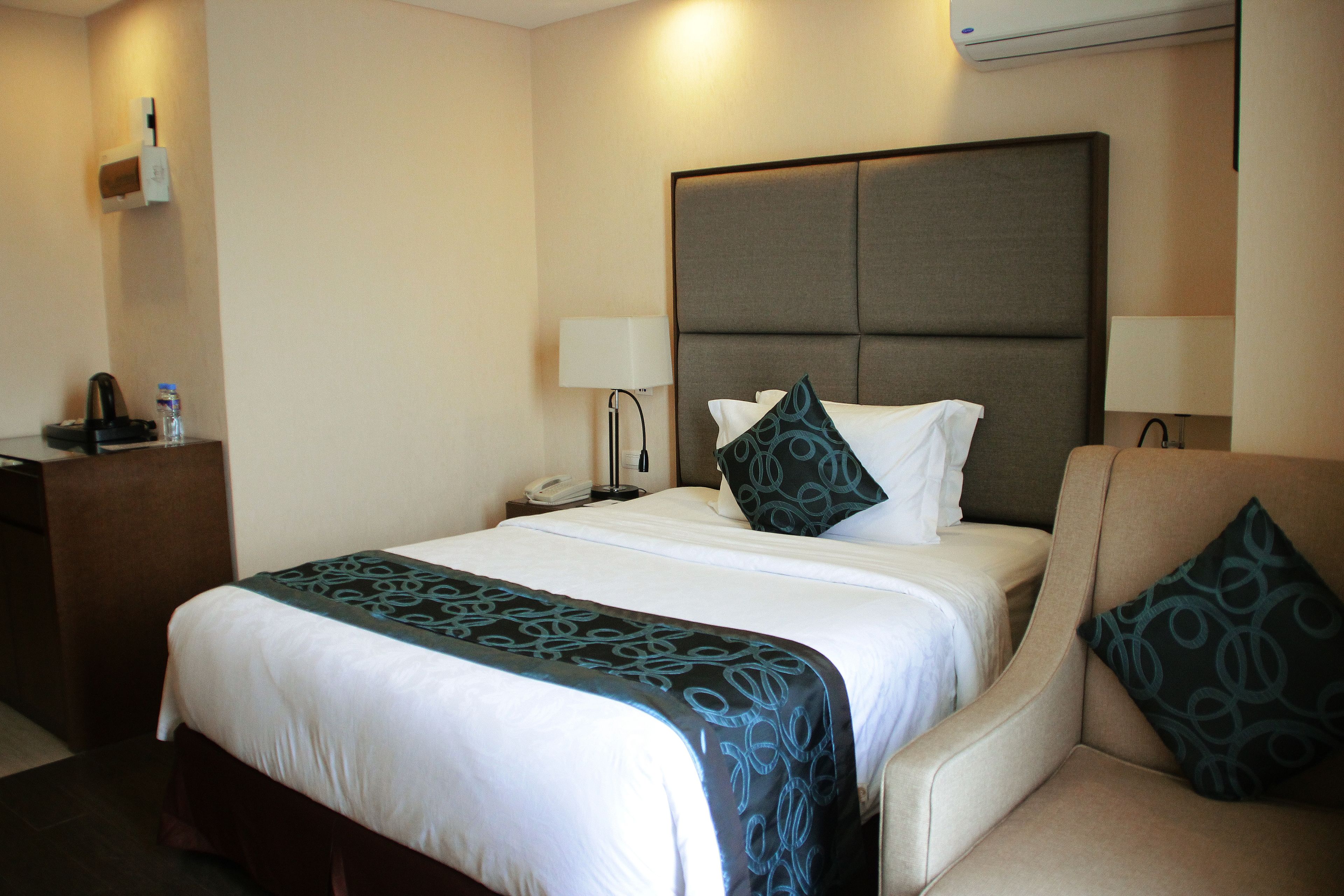 A hotel room with a large bed, a bedside table with a phone, a lamp, a chair with a pillow, and a water bottle on the nightstand.