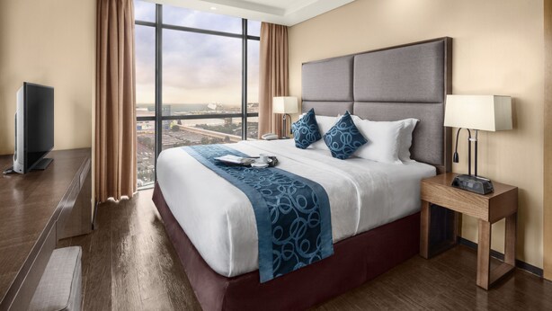 A hotel room with a large bed, a TV, a bedside table with a lamp, and a view of the city. - Golden Phoenix Hotel Manila (Pasay)