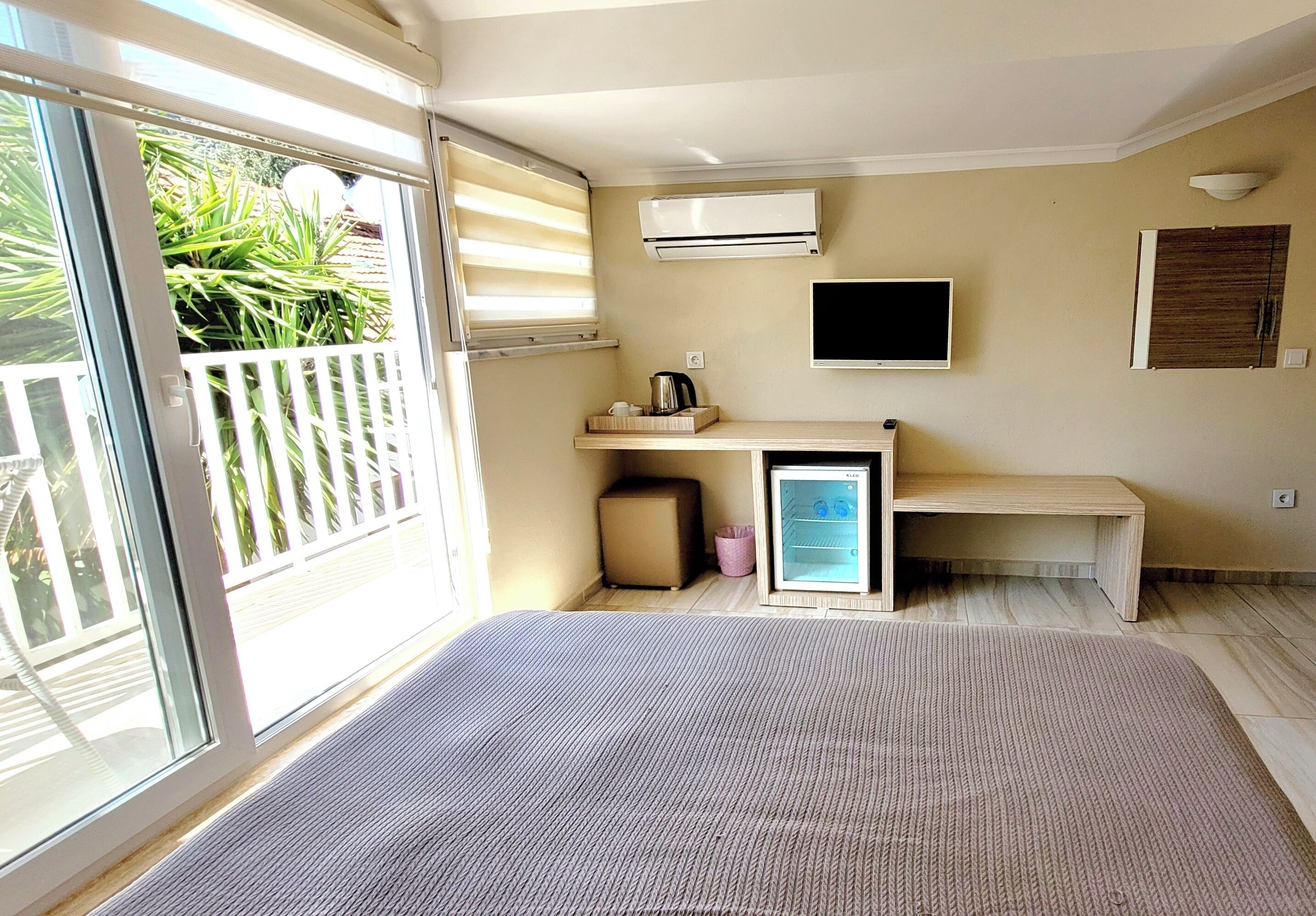 Habitación de hotel con puerta corrediza de vidrio a un balcón, un escritorio pequeño con una silla, un televisor de pantalla plana montado en la pared y un equipo de aire acondicionado integrado.
