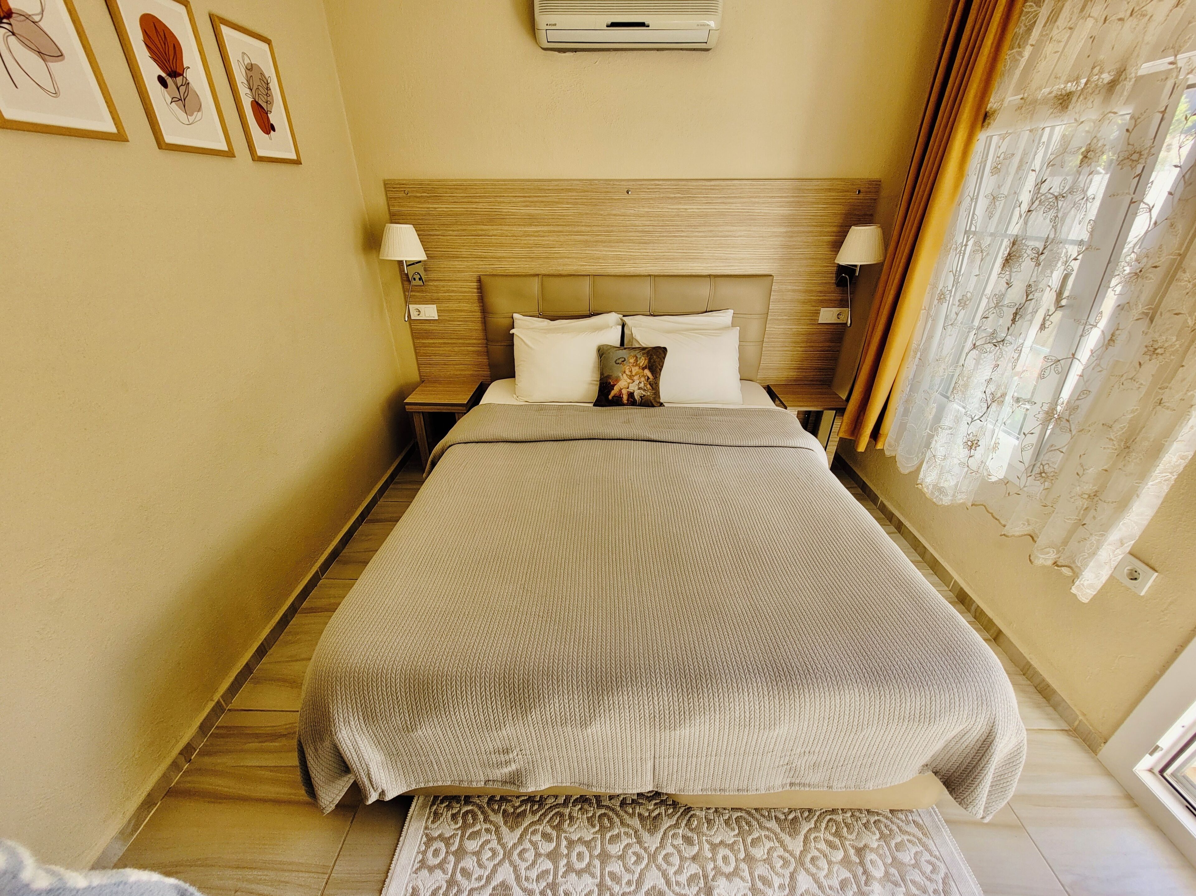A neatly arranged hotel room with a large bed, a wooden headboard, two bedside lamps, a patterned rug, and a window with sheer curtains.