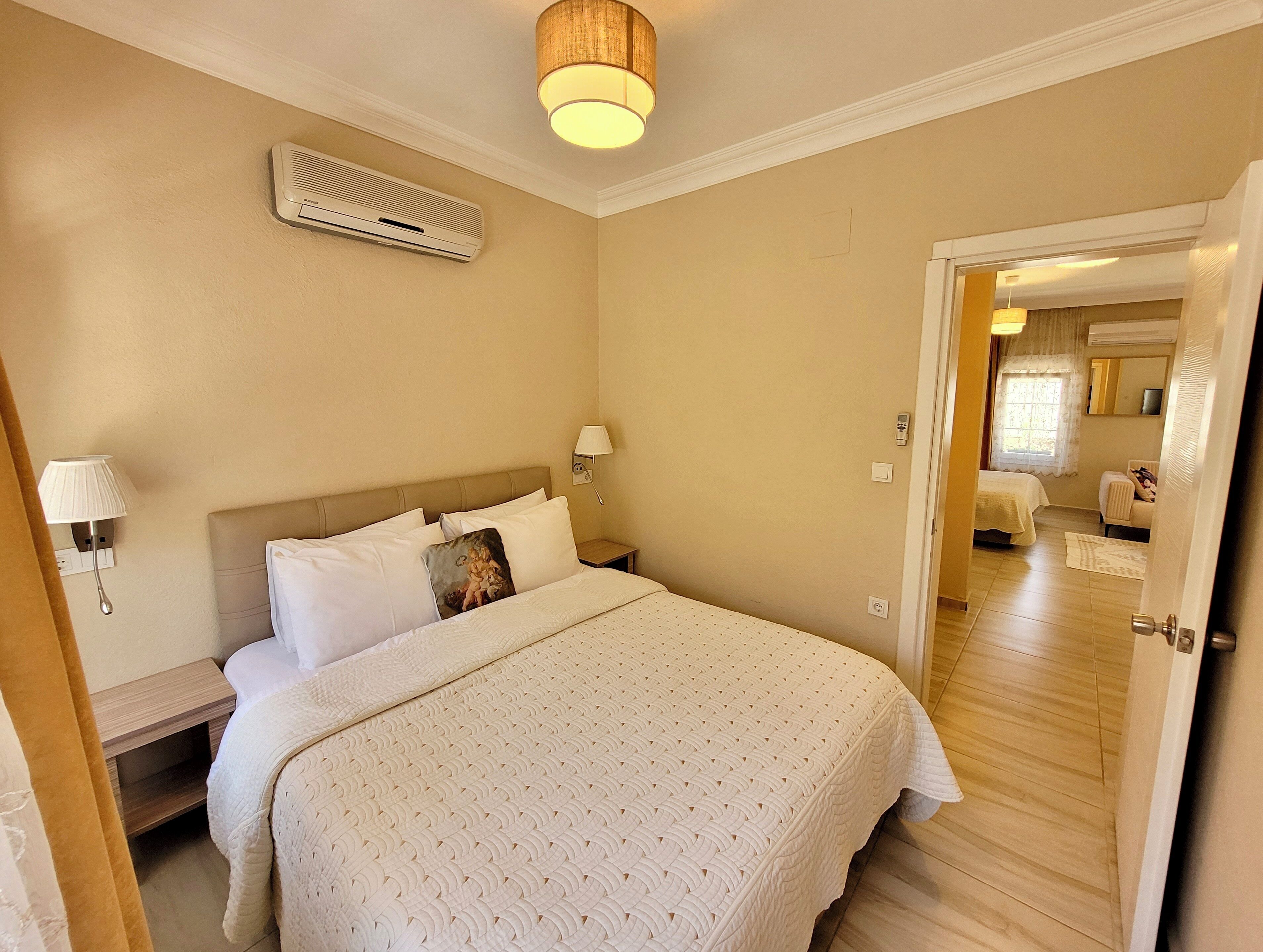 A hotel room with a large bed, bedside lamps, an air conditioning unit, and a door leading to another room.