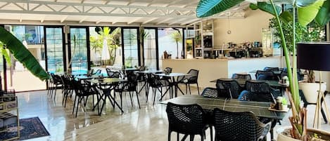 Un espacio interior amplio con varias mesas y sillas, ventanas grandes y plantas en macetas.