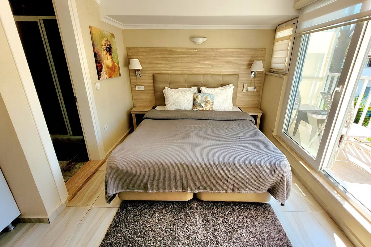 A hotel room with a large bed, two bedside lamps, a painting on the wall, and a view of the outside balcony.