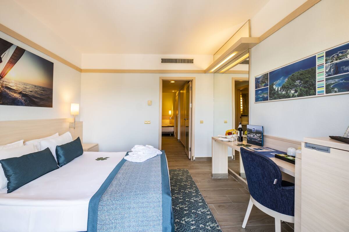 Quarto de hotel com cama, escrivaninha, cadeira e uma janela grande.